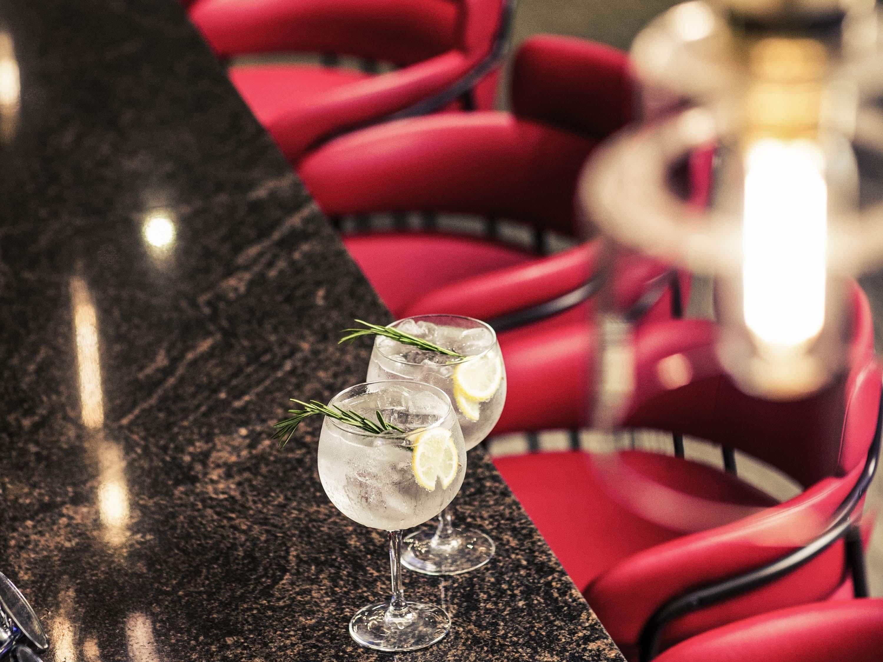 Two glasses of a clear beverage with lemon and rosemary garnish on a marble countertop in a restaurant with red chairs.