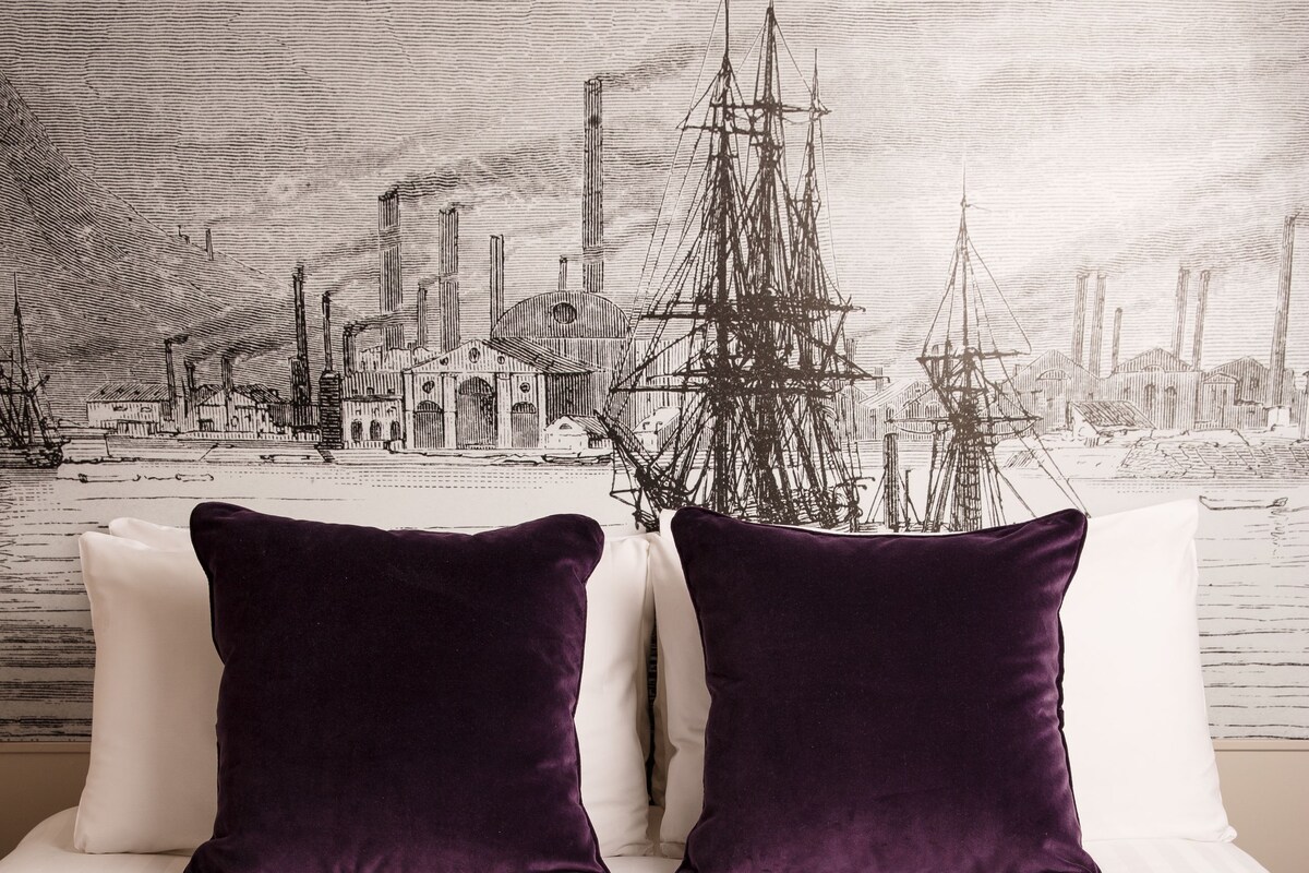A bed with white bedding and two dark purple pillows, a red patterned bedspread, and a cityscape wallpaper in the background.