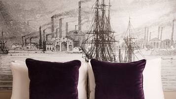 A bed with white bedding and two dark purple pillows, a red patterned bedspread, and a cityscape wallpaper in the background.