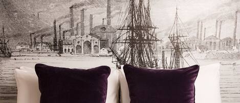 A bed with white bedding and two dark purple pillows, a red patterned bedspread, and a cityscape wallpaper in the background.