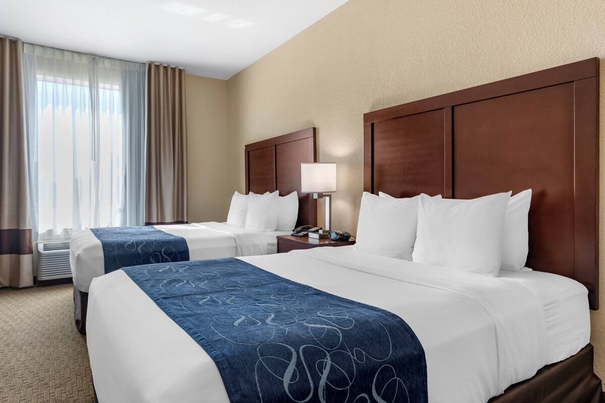 A hotel room with two beds, a nightstand, a lamp, and a window with curtains.