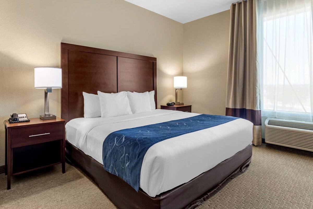 A hotel room with a large bed, two bedside tables with lamps, a desk with a telephone, and an air conditioning unit.