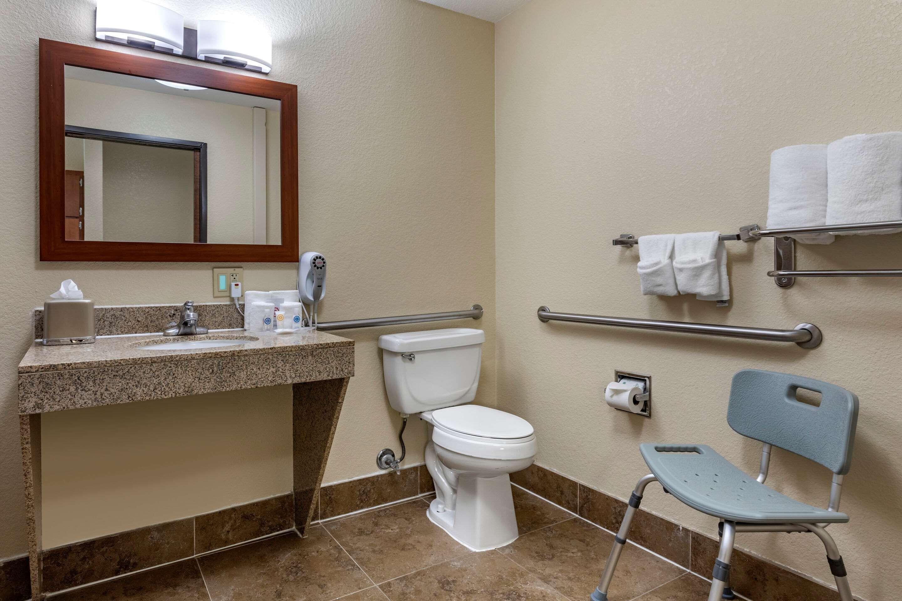 Een badkamer met een wastafel, toilet, spiegel, handdoekhouder en een rolstoeltoegankige stoel.