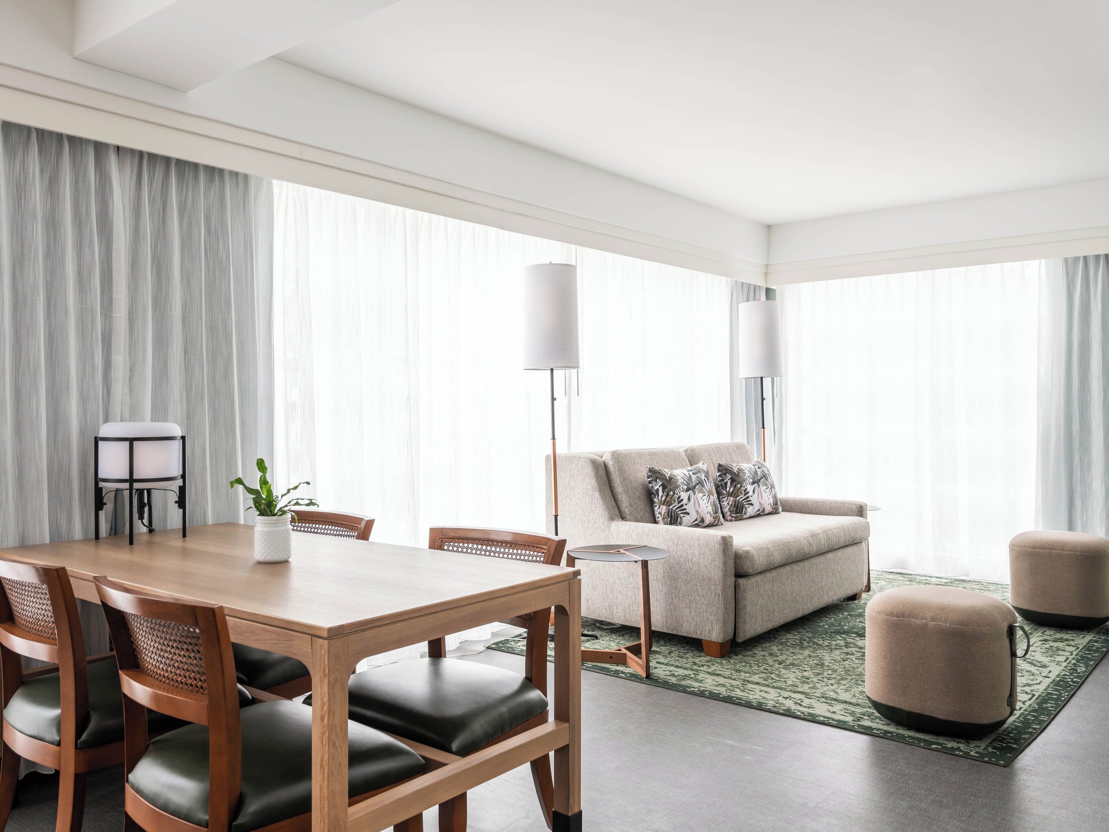 A modern hotel room with a dining area, a sofa, ottomans, and a large window with sheer curtains.