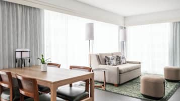 A modern hotel room with a dining area, a sofa, ottomans, and a large window with sheer curtains.