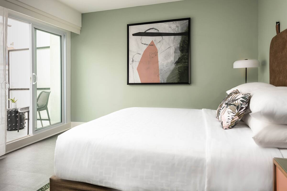 A bedroom with a bed, a framed abstract painting, a nightstand, a lamp, and a view of a balcony with chairs and a plant.