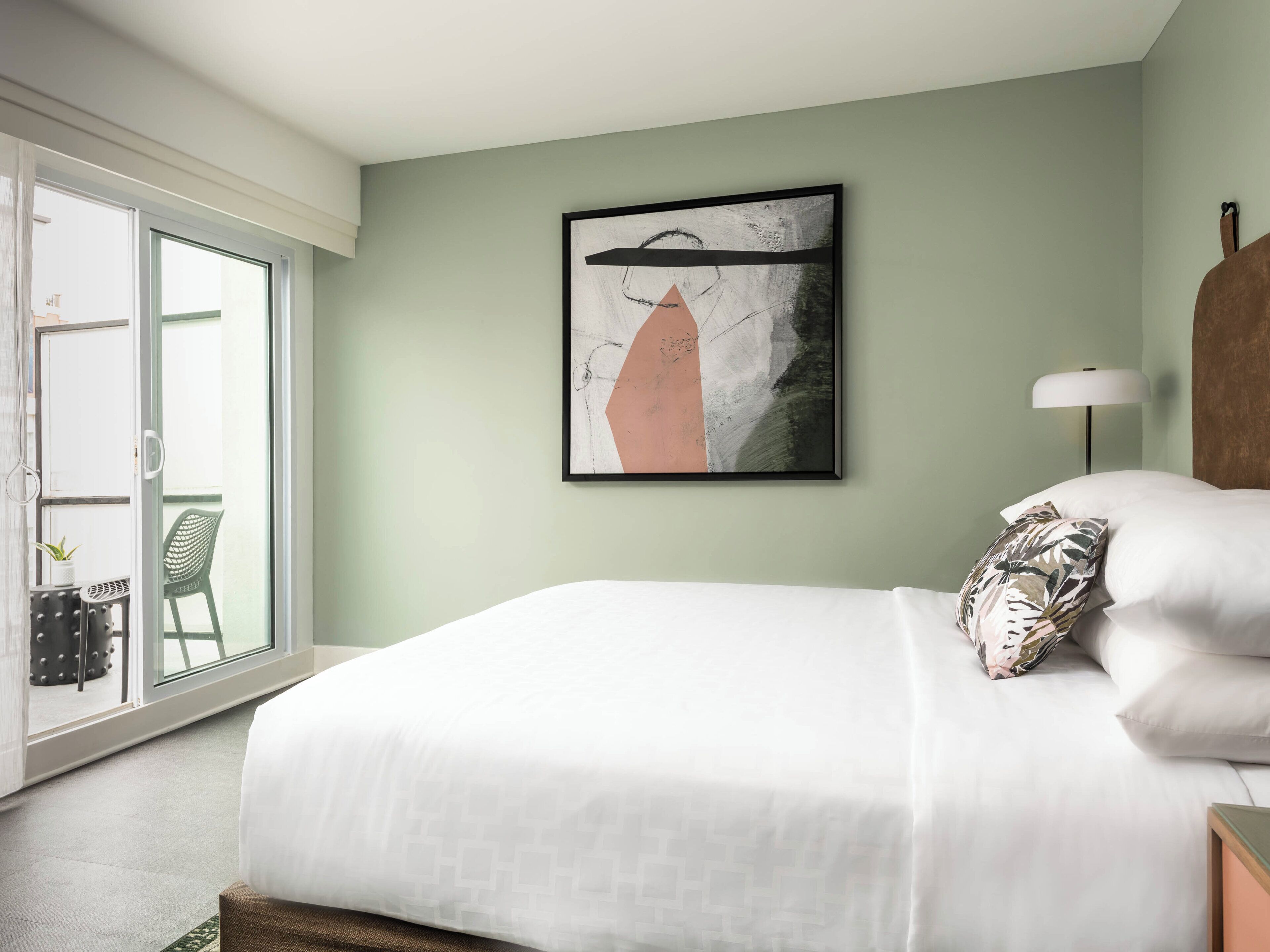 A bedroom with a bed, a framed abstract painting, a nightstand, a lamp, and a view of a balcony with chairs.