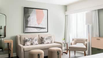 A hotel room with a sofa, ottomans, a chair, a desk, a lamp, and a painting on the wall.