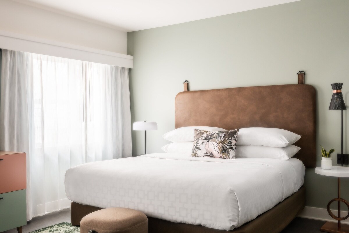 Una cama bien hecha con un almohadón estampado, una lámpara de noche y una pequeña planta en maceta sobre una mesita de noche.