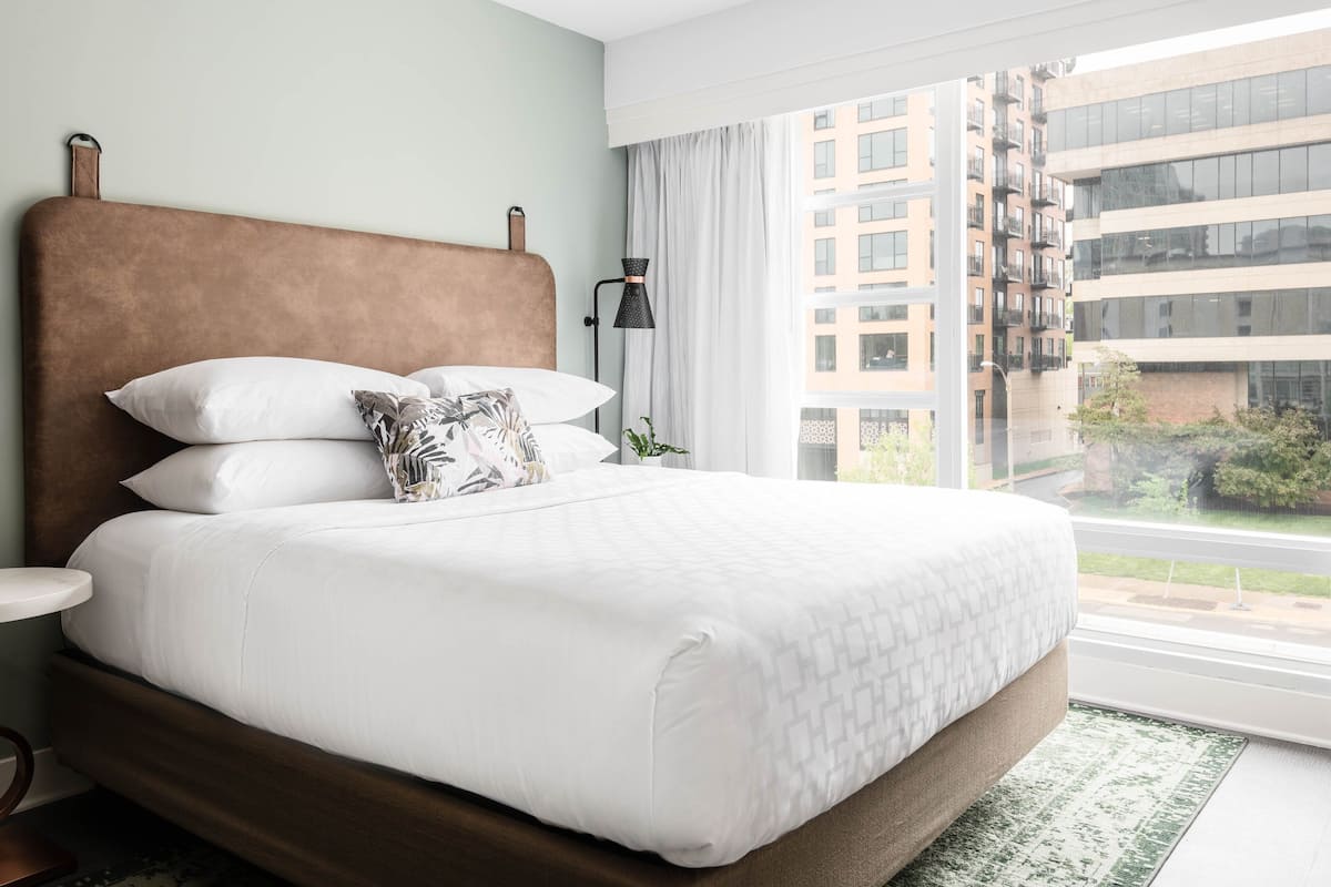 A hotel room with a large bed, a nightstand, a lamp, and a view of the city through large windows.