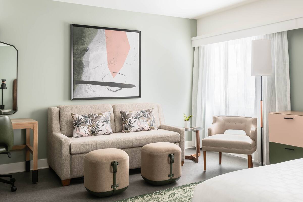A hotel room with a sofa, two ottomans, a chair, a desk, a lamp, and a painting on the wall.