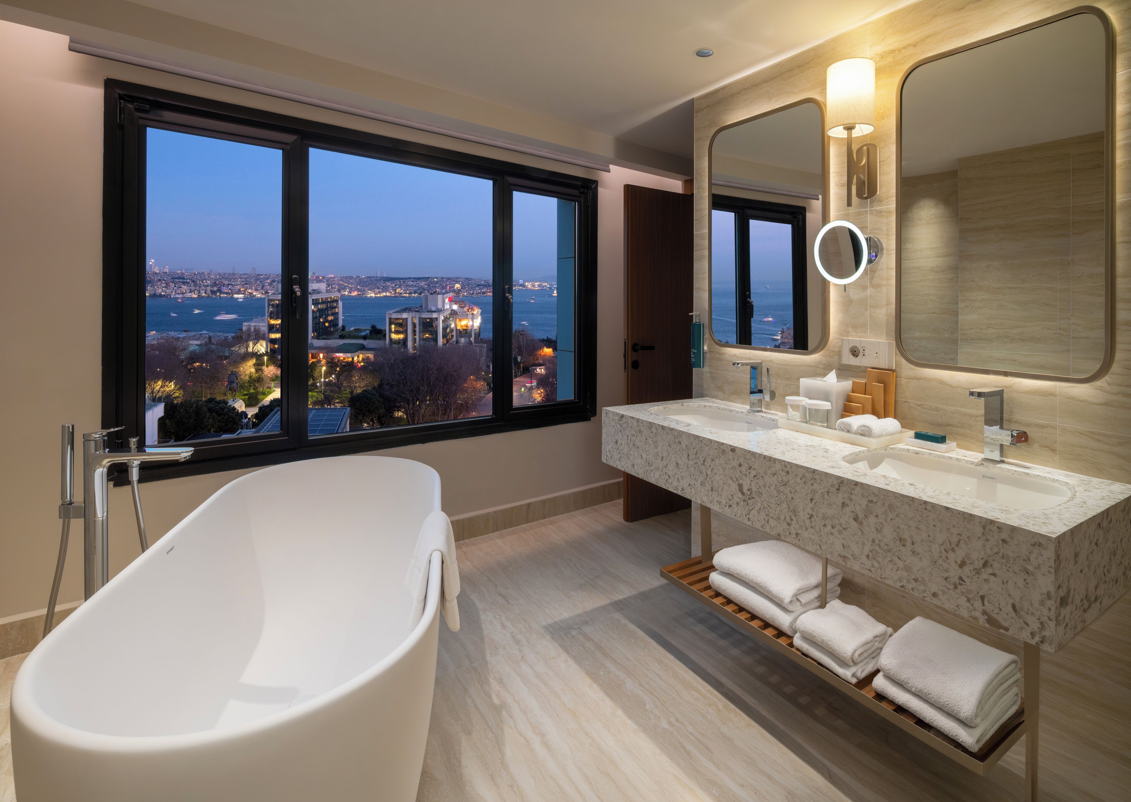 A modern bathroom with a bathtub, a large vanity with a mirror, and a view of a cityscape through large windows.