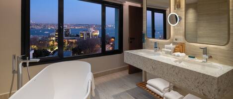 A modern bathroom with a bathtub, a large vanity with a mirror, and a view of a cityscape through large windows.