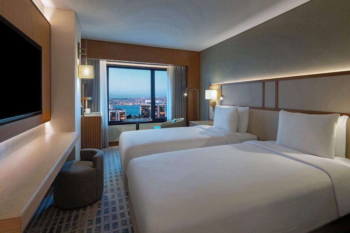 Habitación de hotel con dos camas, televisión, escritorio y ventana grande con vista a la ciudad.