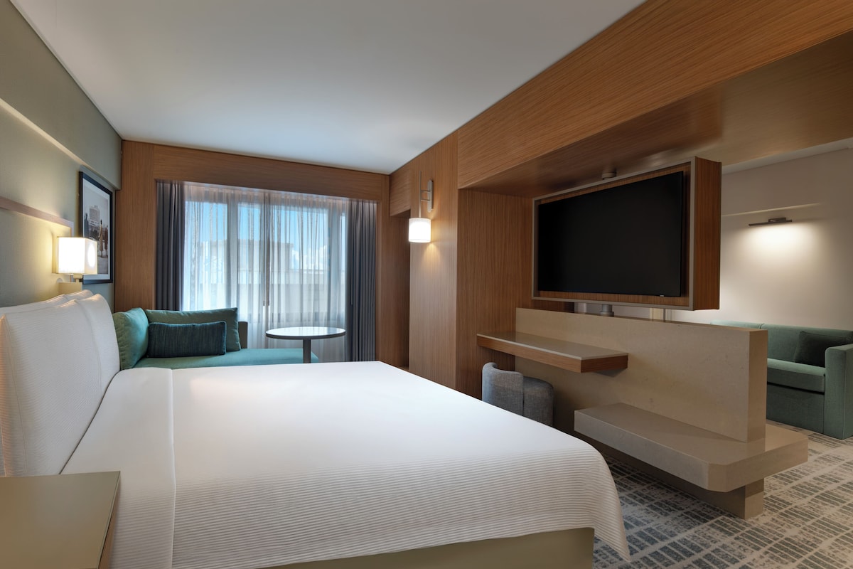 Habitación de hotel con una cama grande, televisión, sofá, mesita y ventana con cortinas.