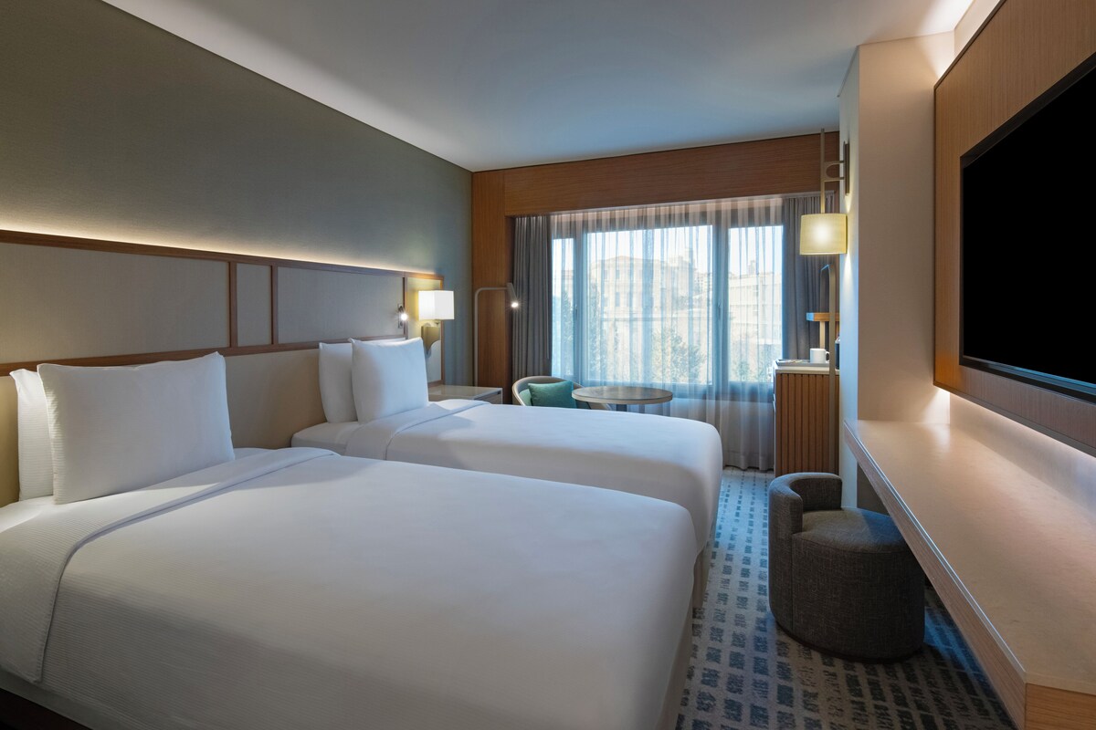 Habitación de hotel con dos camas, televisión, una mesita y ventana con cortinas.