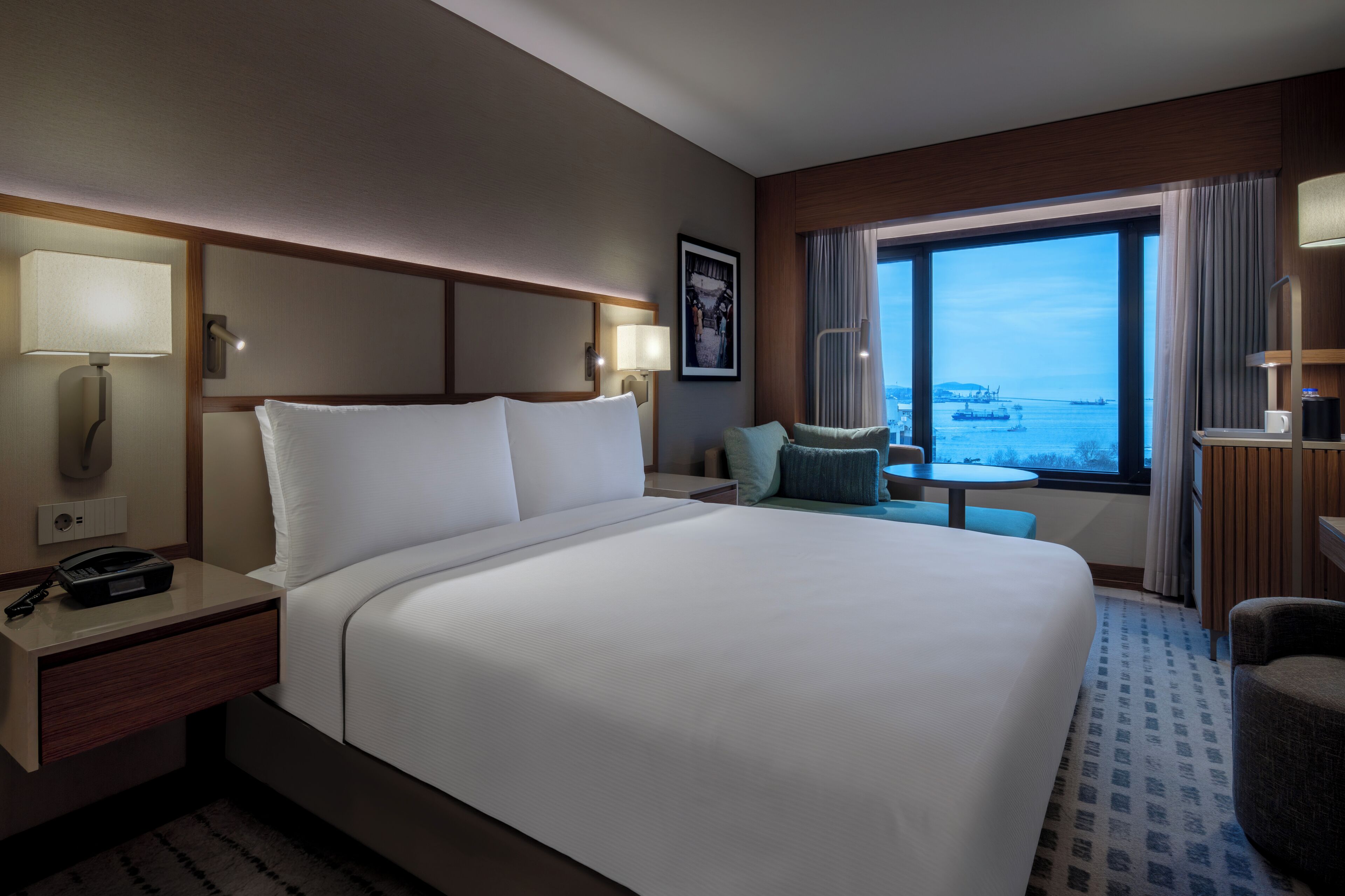 Ein Hotelzimmer mit einem großen Bett, Nachttischlampen, einem Schreibtisch mit Telefon, einem kleinen Tisch mit Stühlen und einem Fenster mit Blick auf die Stadt.