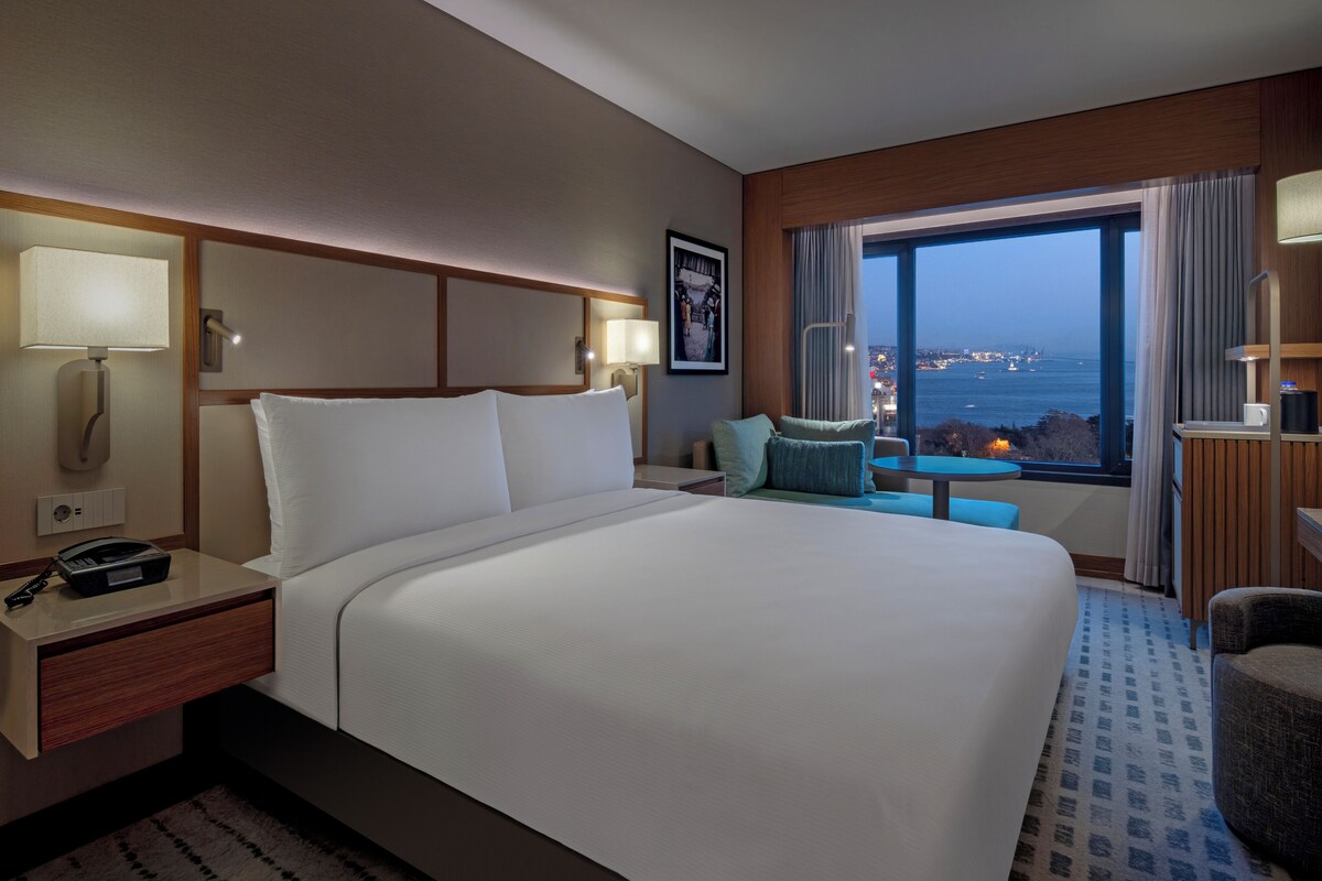 Habitación de hotel con una cama grande, lámparas de noche, un escritorio con teléfono, una mesita con sillas y vistas a la ciudad a través de un ventanal amplio.