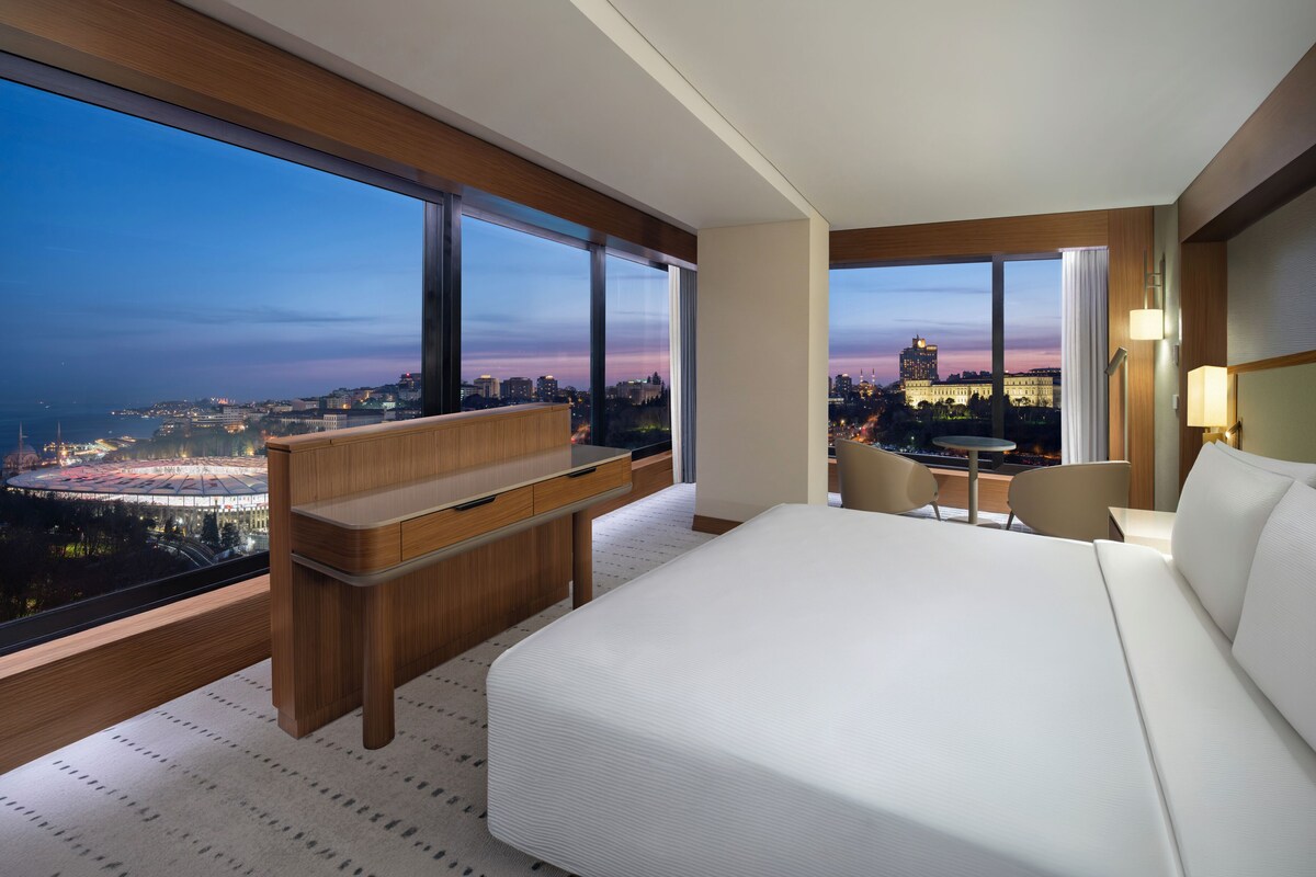 Habitación de hotel con una cama grande, un escritorio, una silla y vistas a una ciudad al atardecer.
