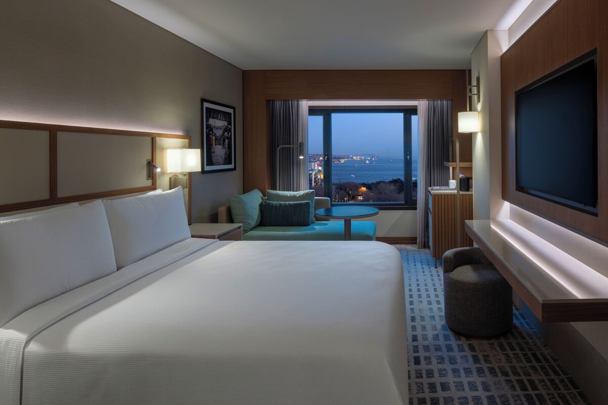 Una habitación de hotel con una cama grande, una zona de estar con vistas, un televisor y un ventanal con vista panorámica de la ciudad.