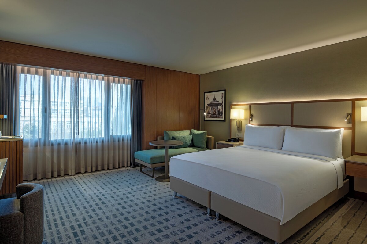 Habitación de hotel con una cama grande, una mesita redonda pequeña, una silla, un escritorio con lámpara y una ventana con cortinas.