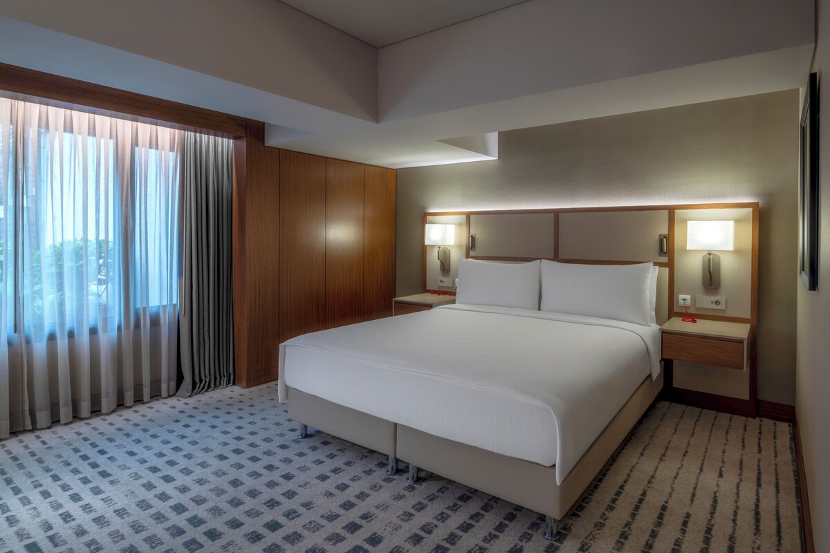 Una habitación de hotel con una cama grande, paneles de madera, dos lámparas de noche y una ventana con cortinas traslúcidas.