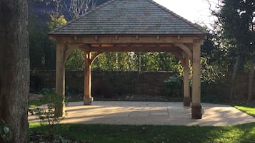 A gazebo with a tiled roof and wooden beams, surrounded by a grassy area and trees.