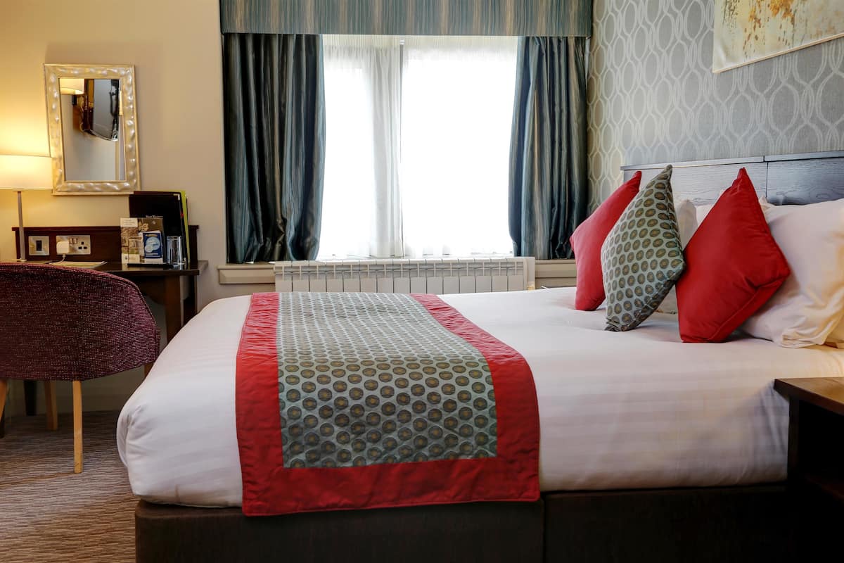 Ein Hotelzimmer mit einem großen Bett, einem beleuchteten Spiegel, einem Schreibtisch und einem Fenster mit Vorhängen.