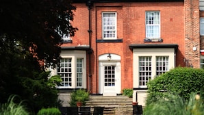 A traditional brick building with a symmetrical facade, a central entrance with a white door, and a small front garden with a bench and potted plants.