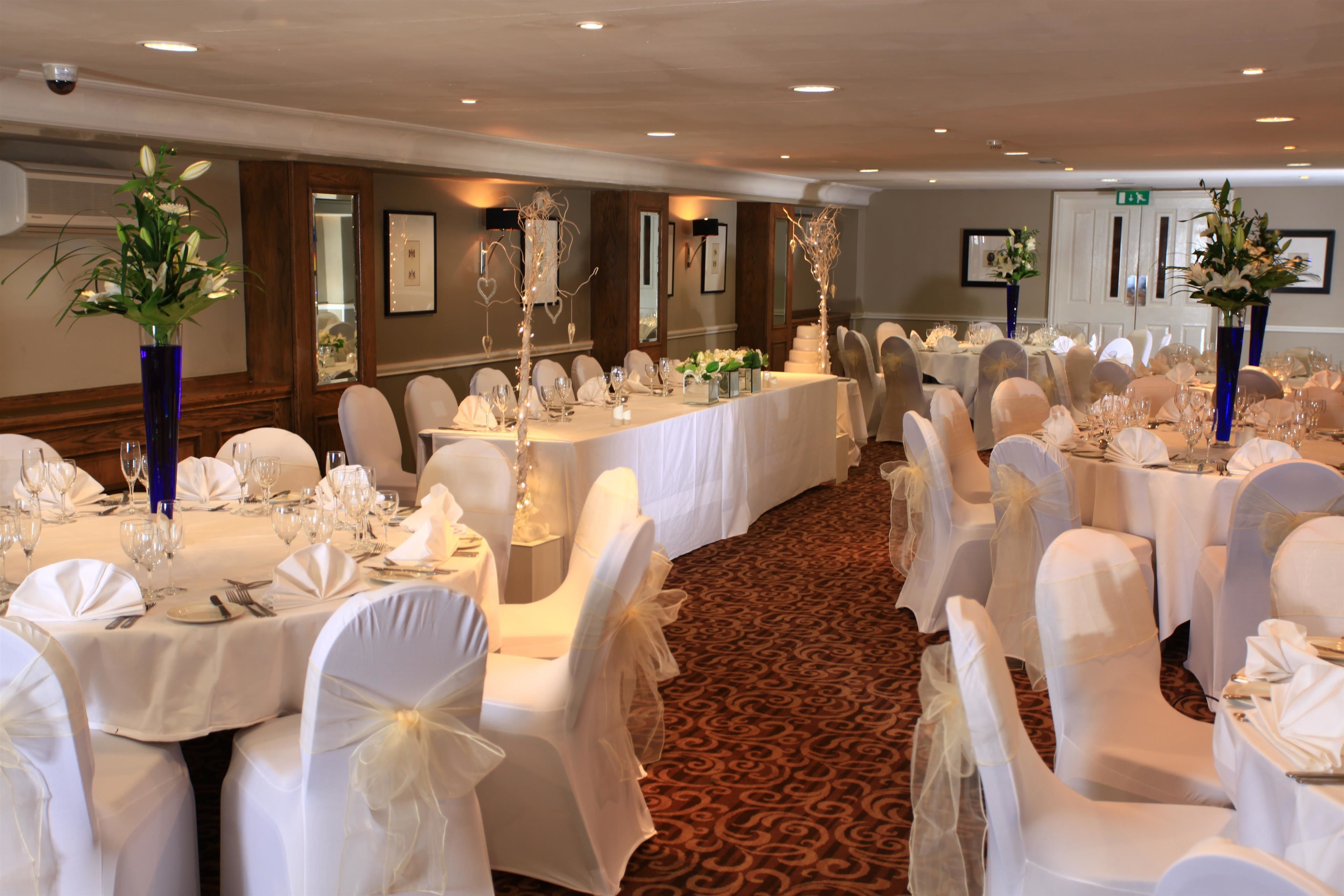 Une salle de banquet avec des tables rondes dressées pour le repas, des chaises blanches et de grands vases remplis de fleurs.