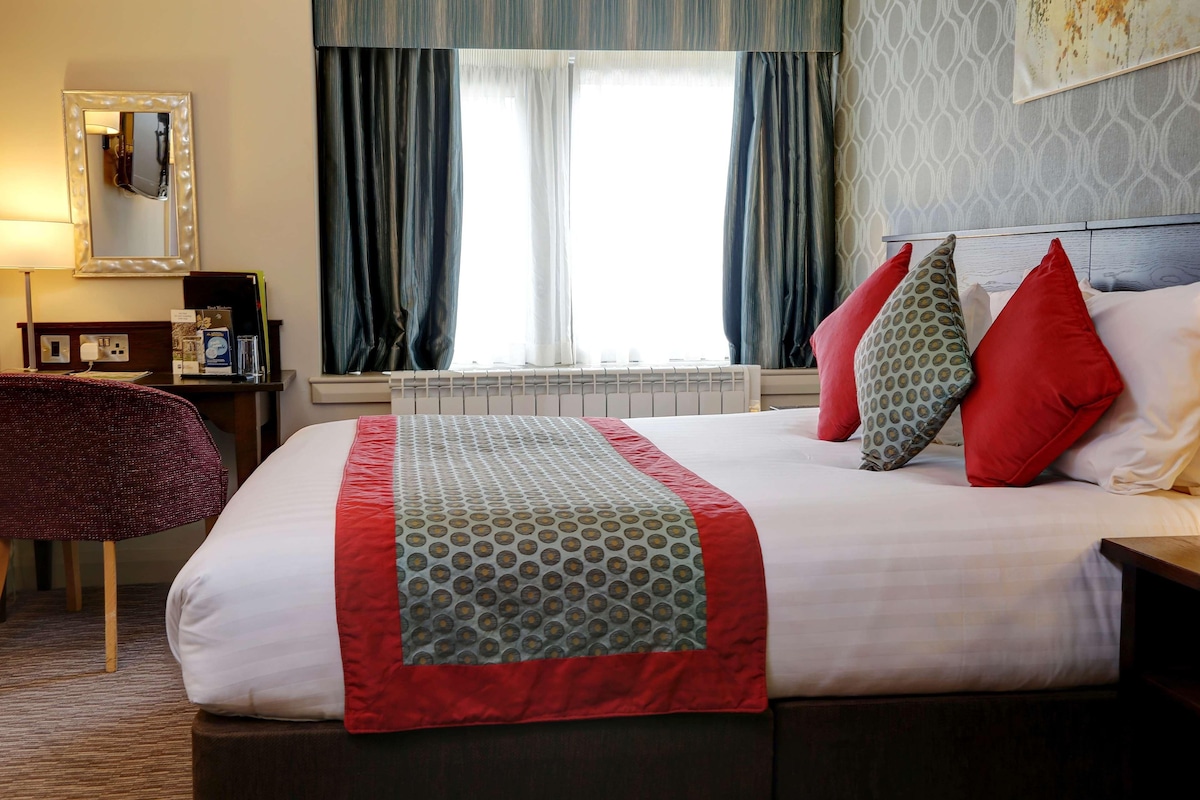 Een hotelkamer met een groot bed, een spiegel met verlichting, een raam met gordijnen en een bureau met lamp.