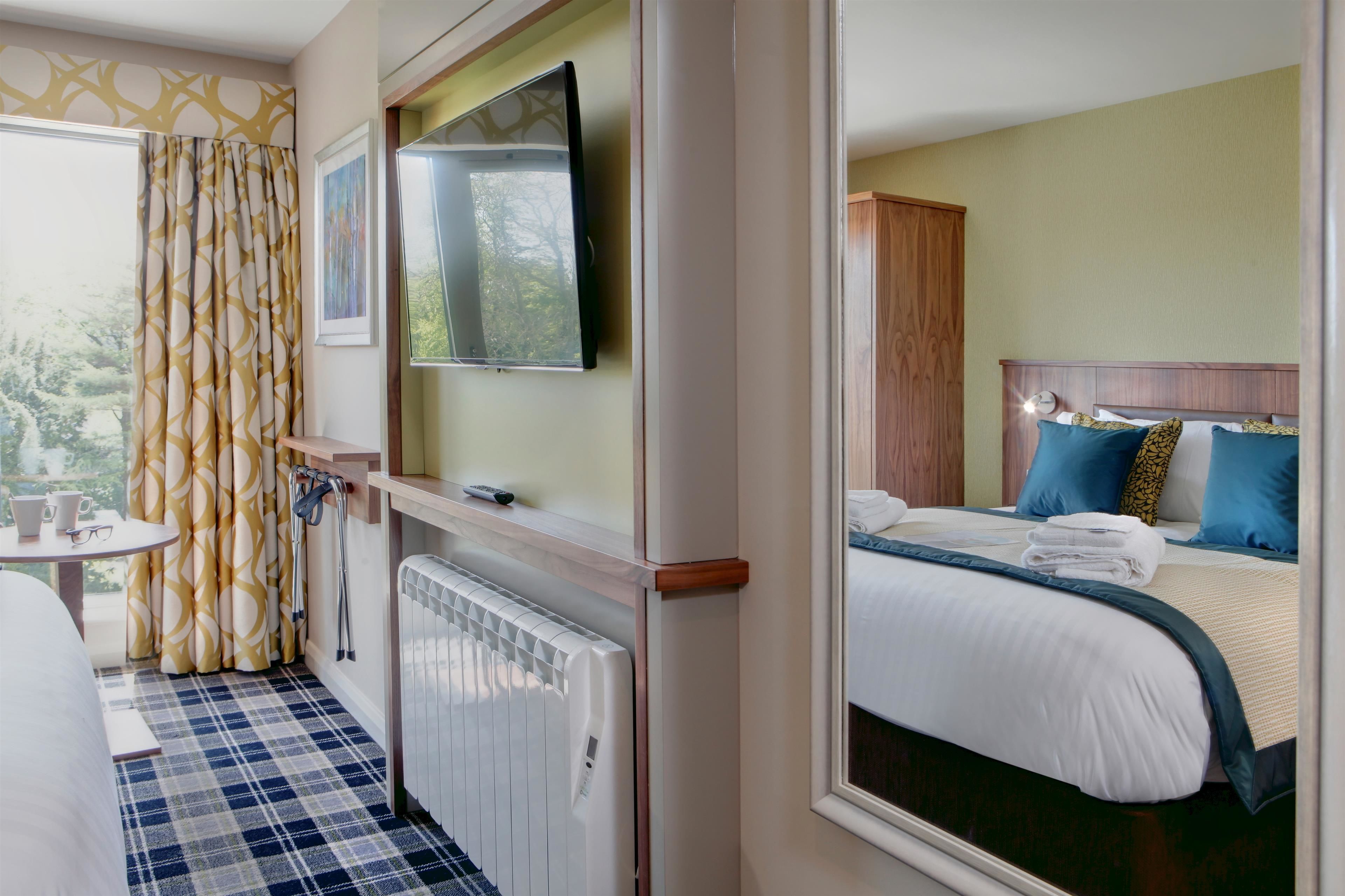 Quarto de hotel com uma cama, televisão presa na parede, uma mesinha com uma chávena e copos, e uma janela com cortinas.