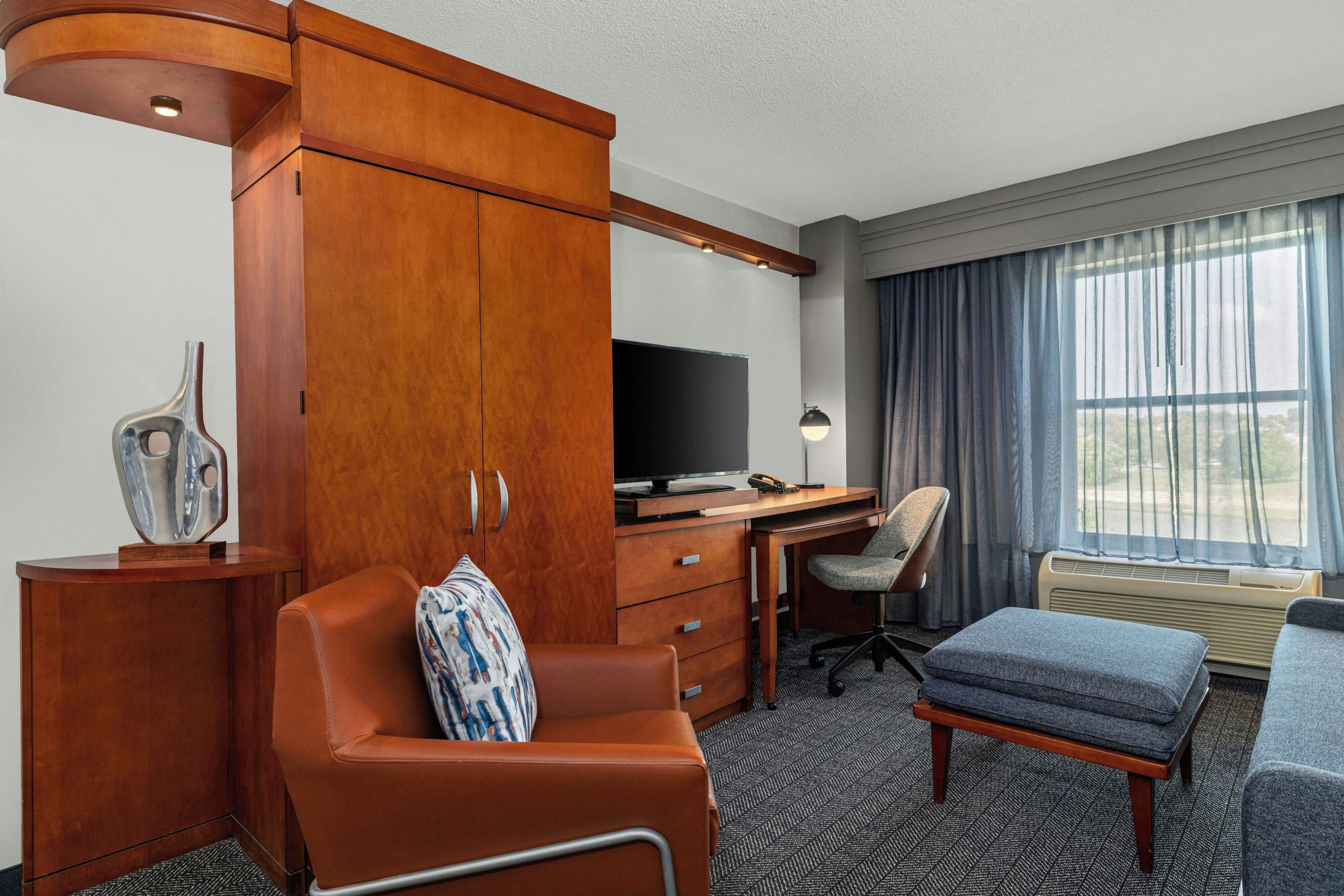 A hotel room with a wooden wardrobe, a desk with a computer, a television, a chair, an ottoman, and a large window with curtains.