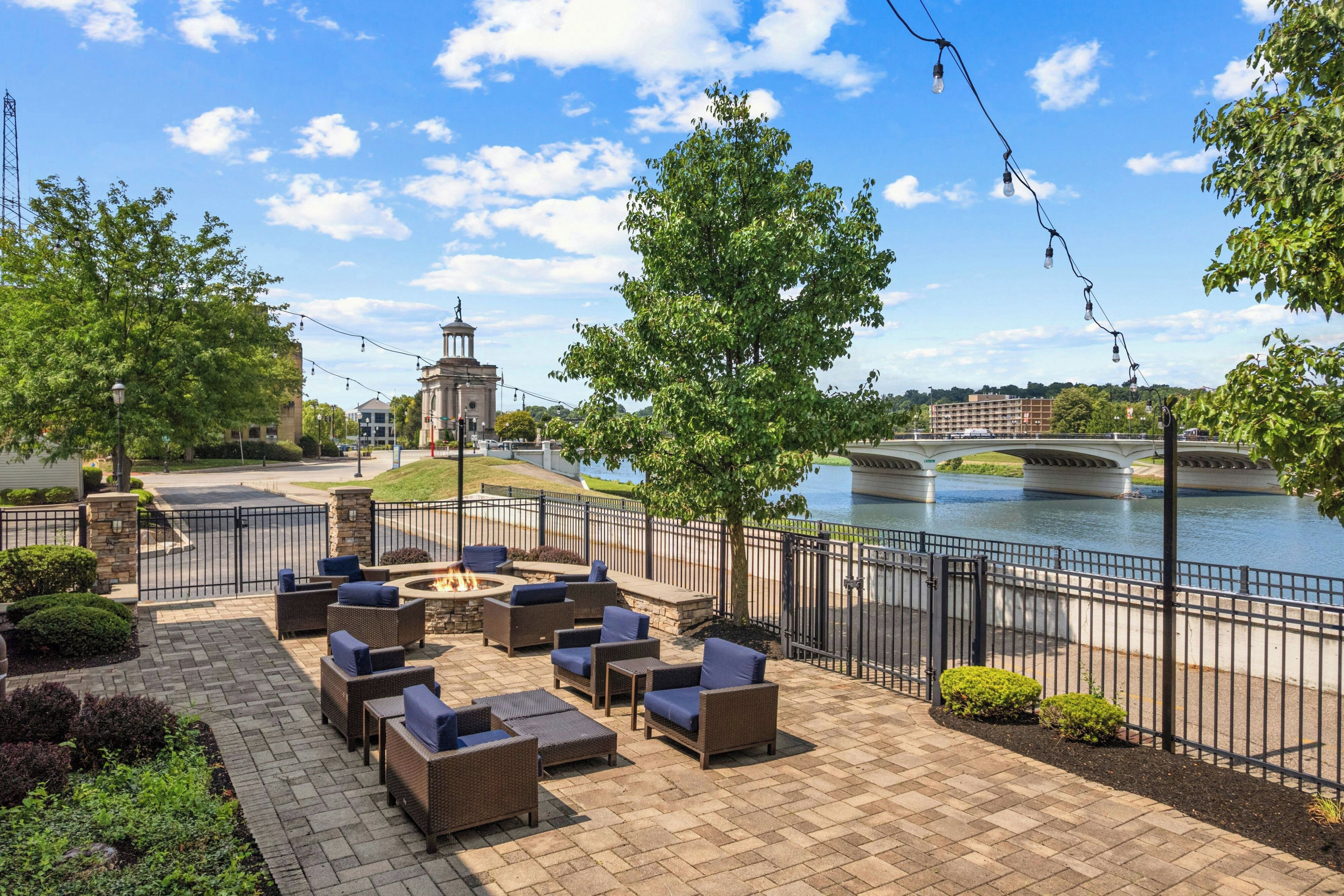 An outdoor patio with wicker furniture and a fire pit, overlooking a river and a bridge.