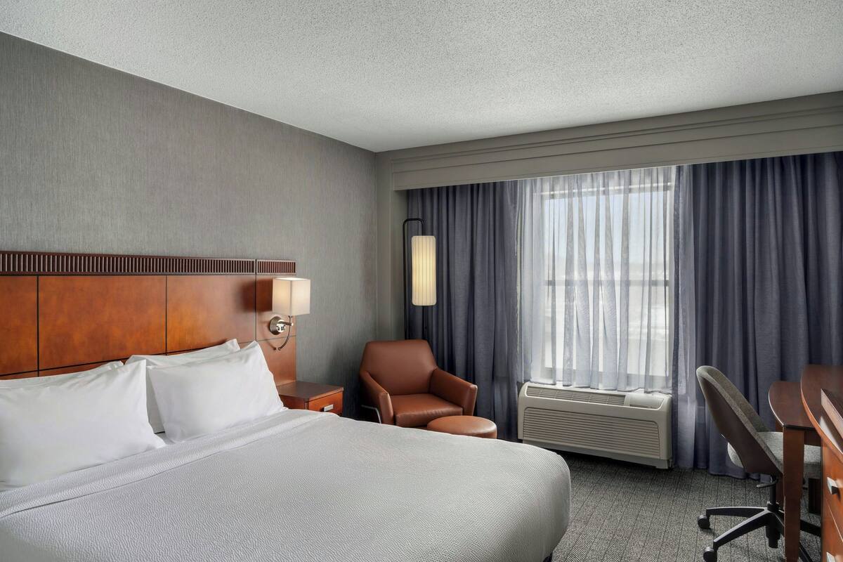 A hotel room with a large bed, a desk, a chair, a lamp, and a window with curtains.