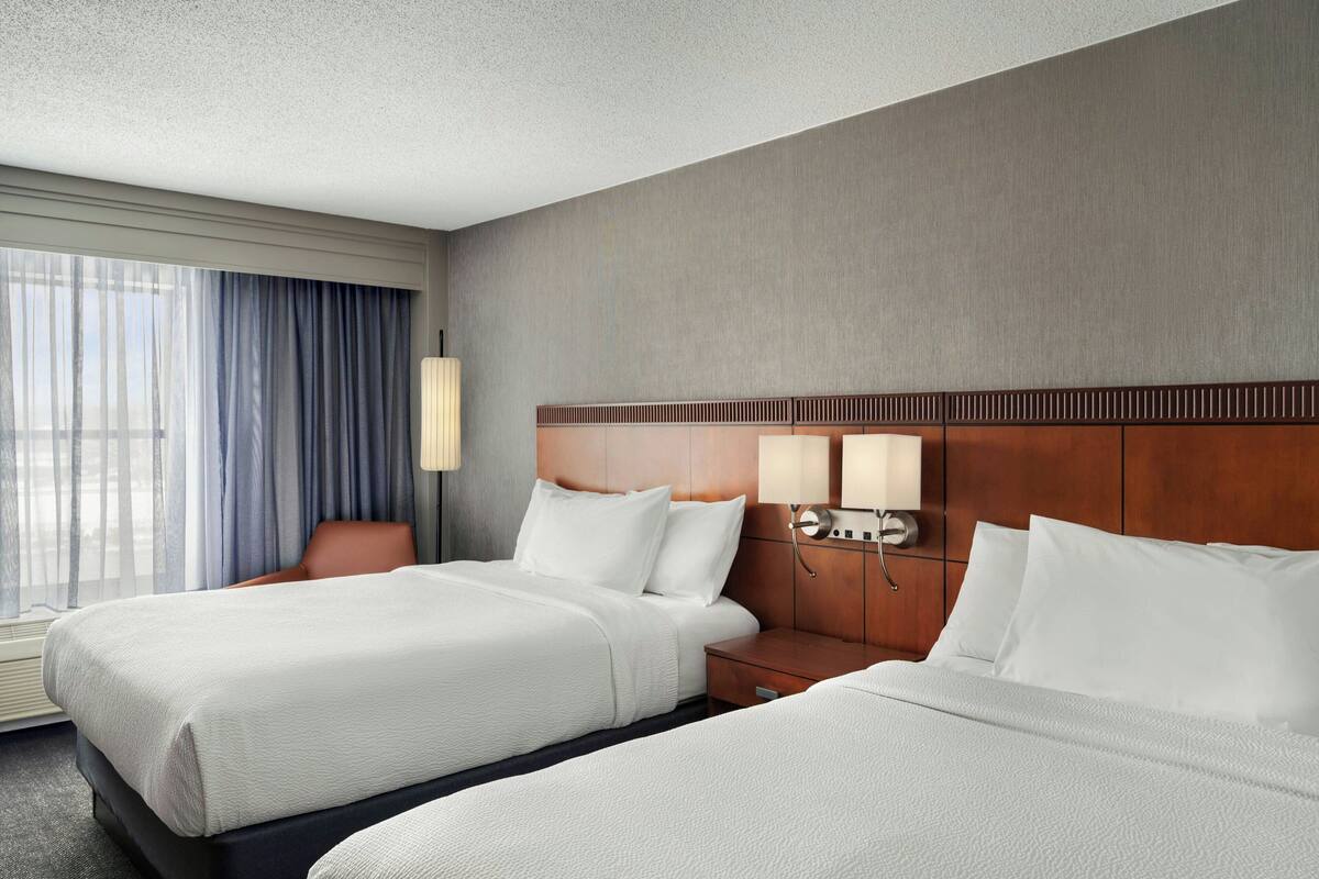 A hotel room with two beds, a wooden headboard, bedside lamps, a chair, and a large window with curtains.