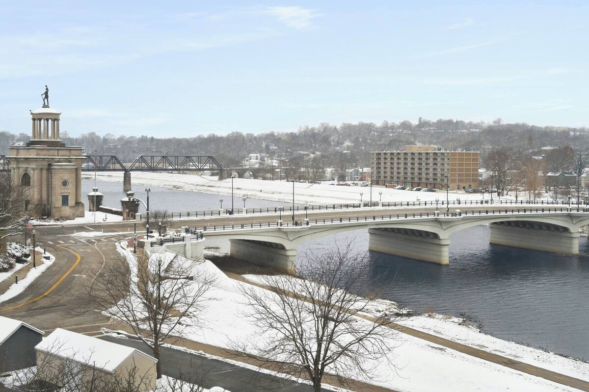 A bridge over a river with a statue on the left, surrounded by snow-covered ground and buildings.