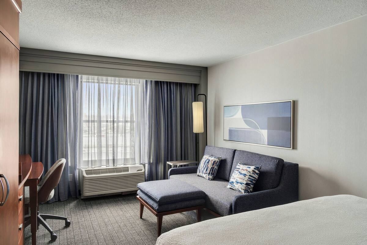 A hotel room with a bed, a sofa, a desk, and a chair. There is a window with curtains, a wall-mounted lamp, and a framed picture.