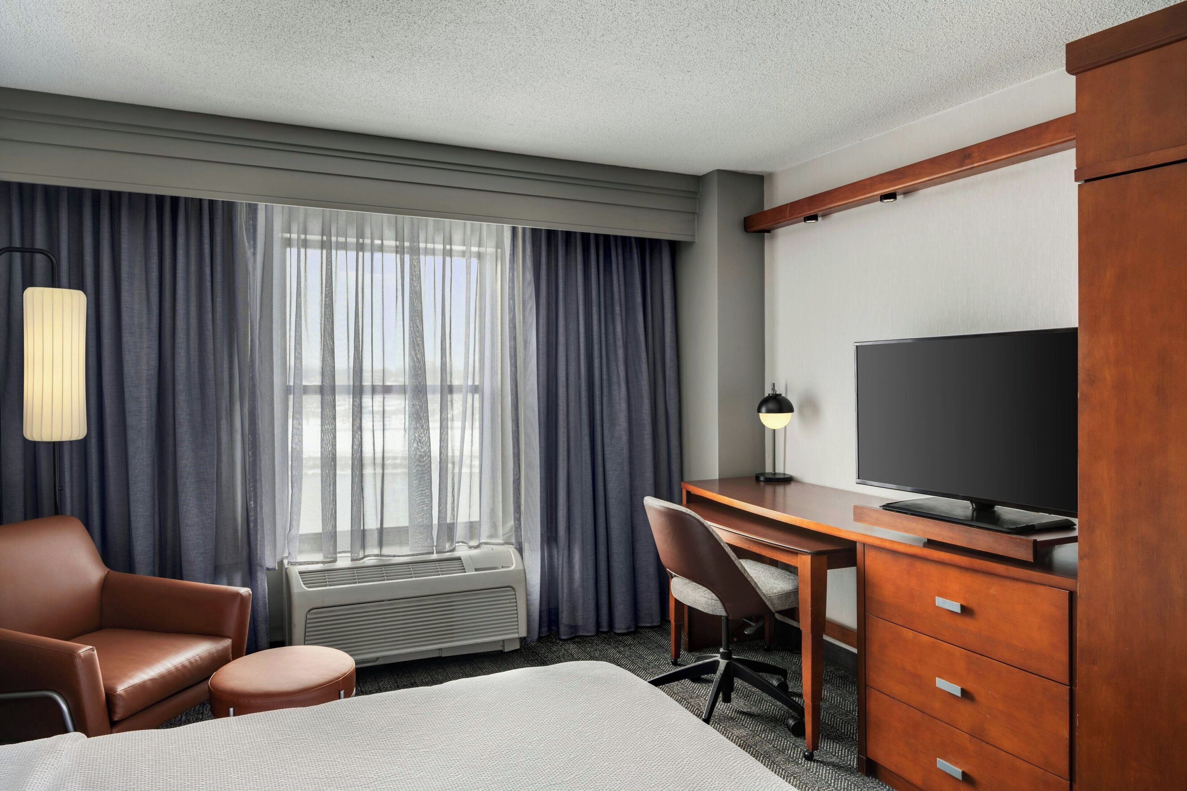 Habitación de hotel con cama, escritorio, televisión, silla y un amplio ventanal con cortinas.