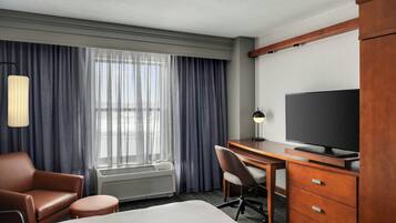 Habitación de hotel con cama, escritorio, televisión, silla y un amplio ventanal con cortinas.