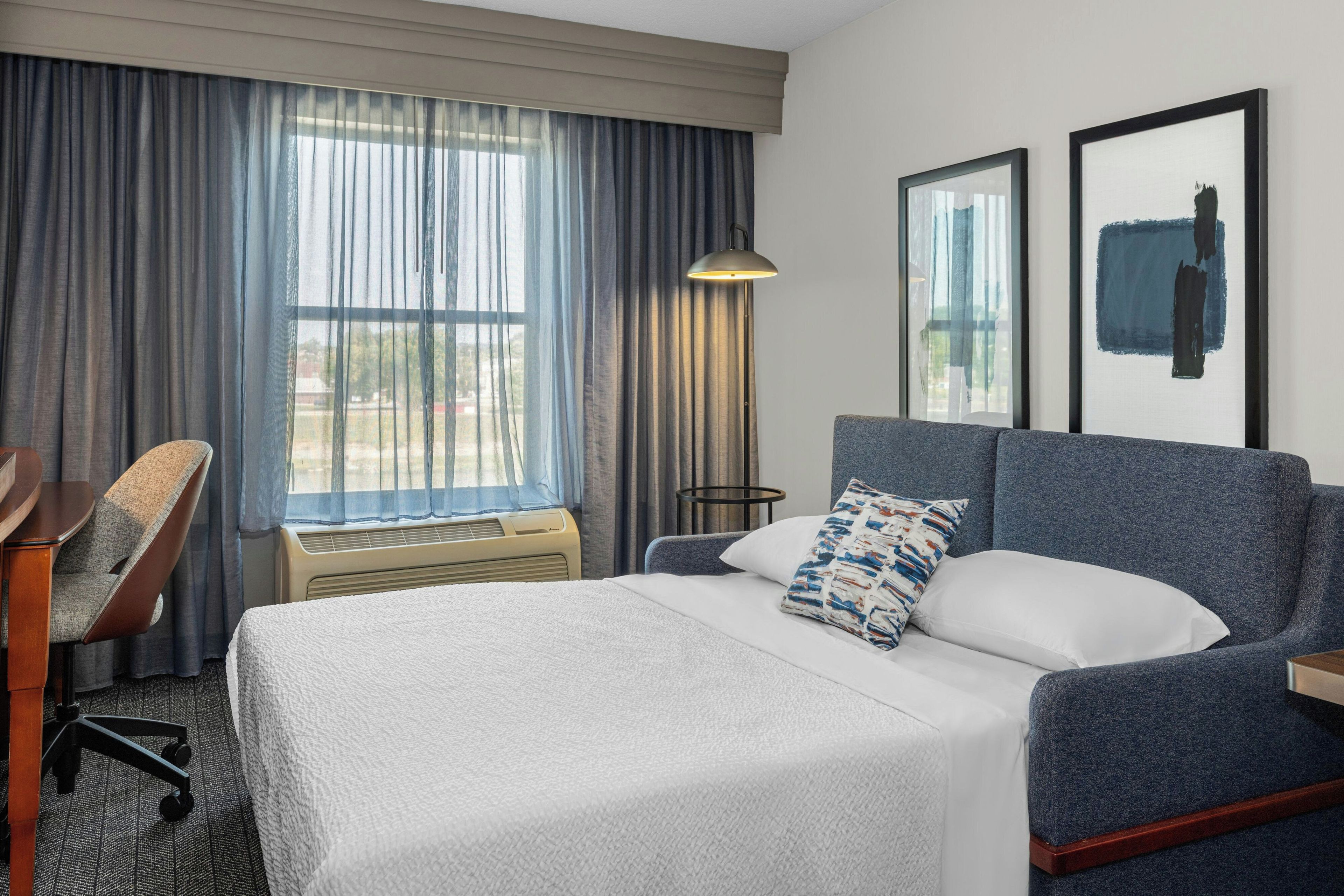 A hotel room with a bed, a desk, a chair, a lamp, and two framed pictures on the wall.