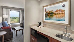 A hotel room with a kitchenette, a dining area with a table and chairs, a large window with a view of a park, and a framed picture of a lake. - Hampton Inn & Suites Alexandria Old Town Area South (Alexandria)