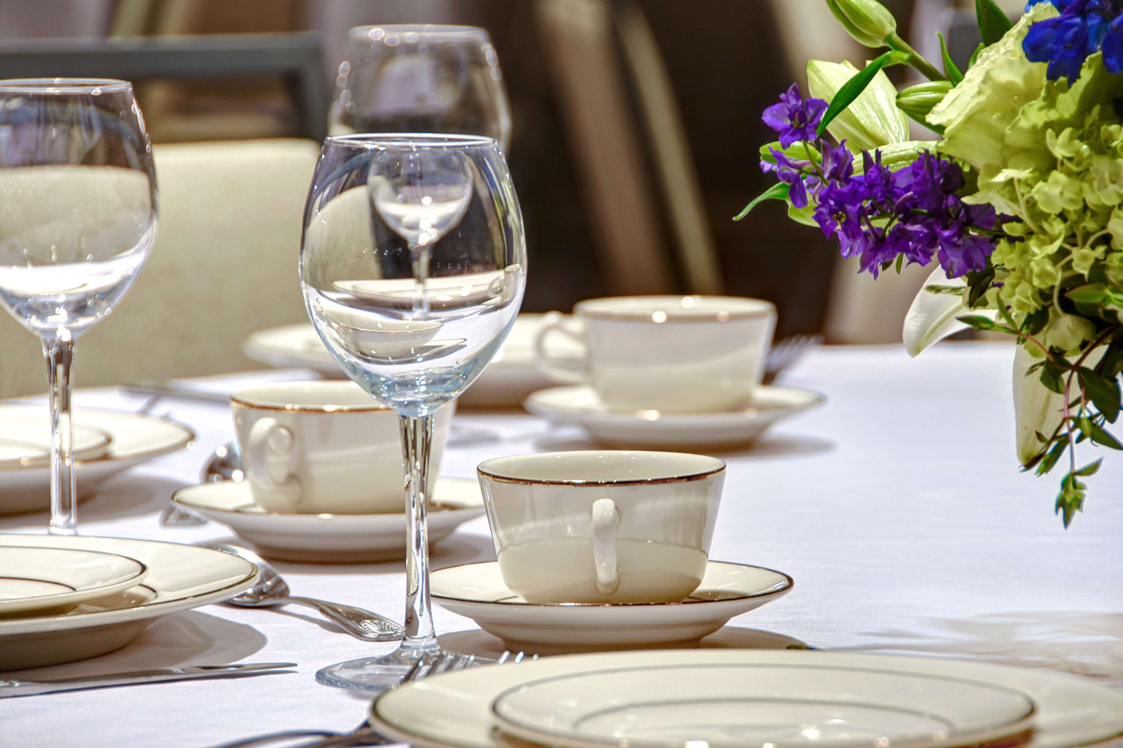 A table set with white plates, cups, and a wine glass, with a vase of flowers on the right.