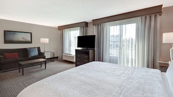Habitación de hotel con una cama grande, televisión de pantalla plana, sofá, mesa de centro y vistas a la ciudad desde las ventanas.