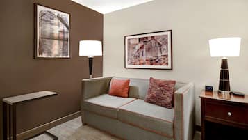 Habitación de hotel con un sofá gris, dos cuadros en la pared, una mesita de noche con lámpara y una pequeña mesita auxiliar de madera.