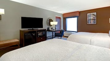 A hotel room with two beds, a television, a desk with a lamp, a chair, and a window with blinds.