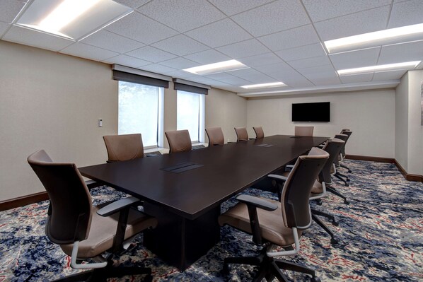 Un salón de conferencias con una larga mesa, varias sillas, un televisor de pantalla plana y ventanas amplias.