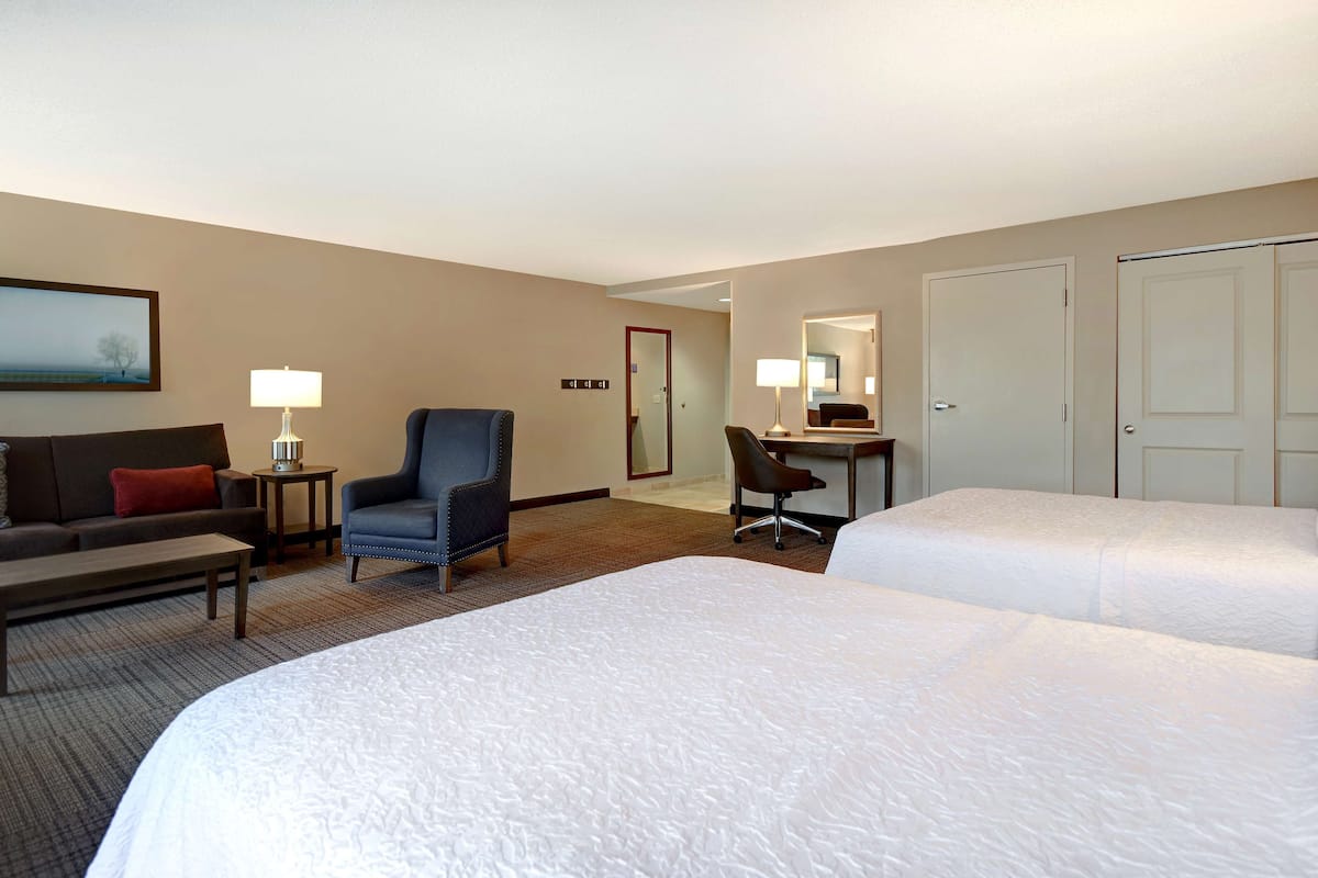A hotel room with two beds, a desk, a chair, a sofa, a lamp, and a painting on the wall.
