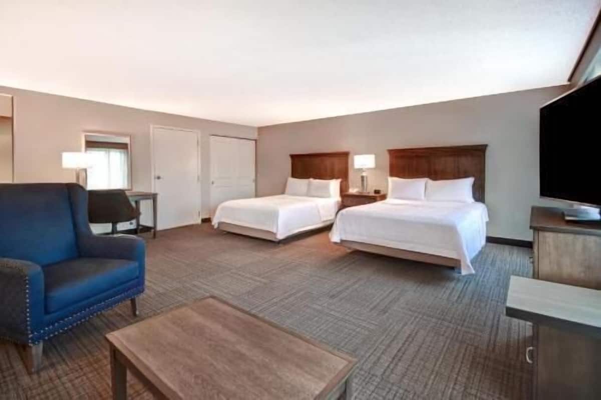 A hotel room with two beds, a blue armchair, a wooden coffee table, and a TV.