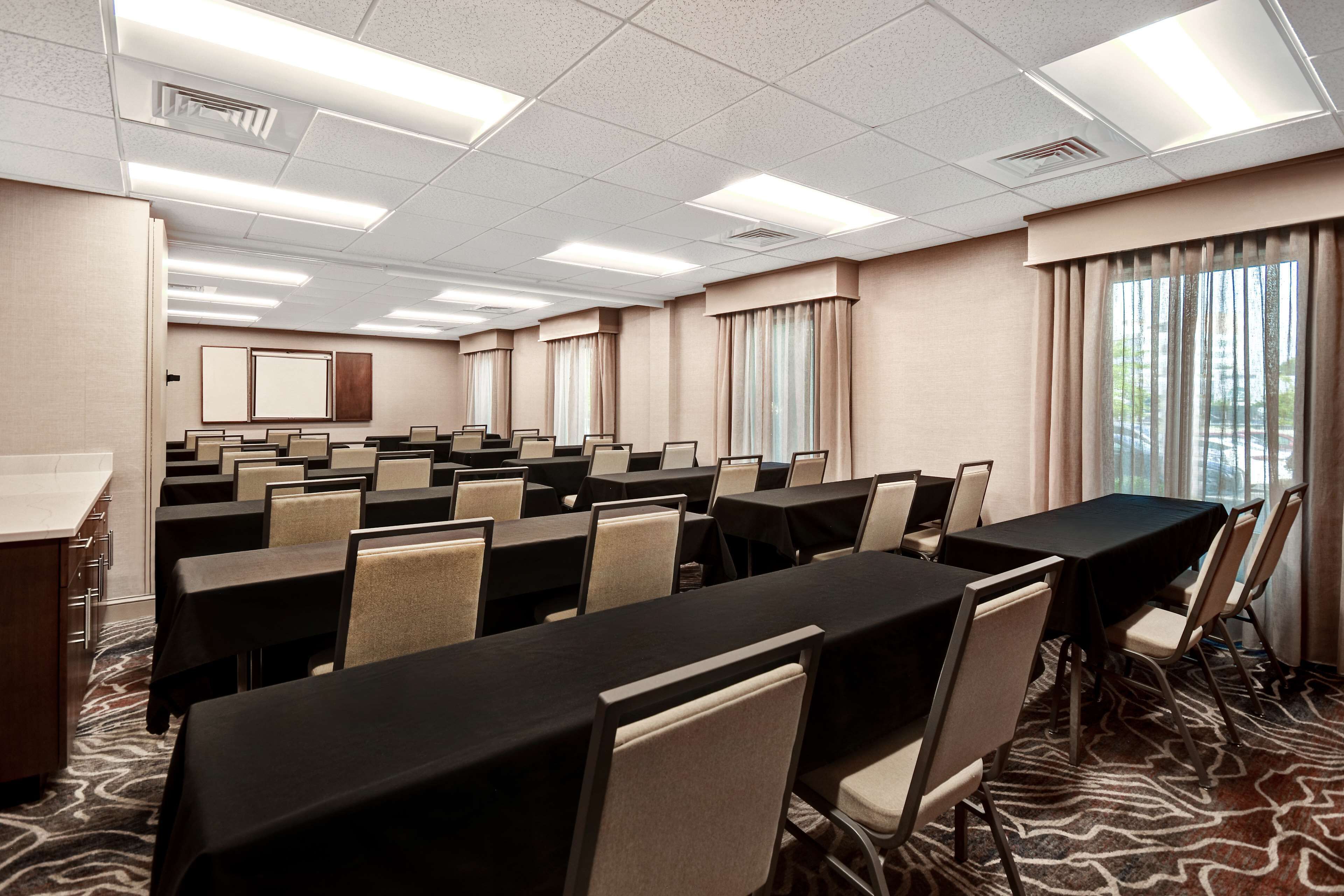 Une salle de conférence équipée de plusieurs tables et chaises, d’un pupitre et de grandes fenêtres avec des rideaux.